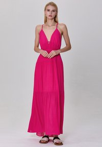 Robe maxi sans manches rose avec un décolleté en V profond, taille froncée et jupe à volants. Tissu léger avec un design fluide. Sandales plates.