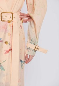Trench coat a fantasia pesca con design floreali, maniche lunghe e vita aderente. Caratterizzato da accessori in tonalità oro e una texture di tessuto morbido.