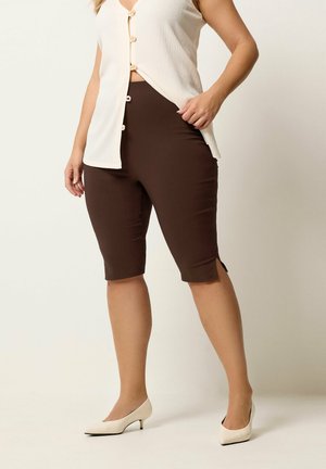 Frau trägt ein weißes ärmelloses, geknöpftes Oberteil, braune, knielange, figurbetonte Shorts und weiße flache Schuhe, steht vor einem schlichten Hintergrund.