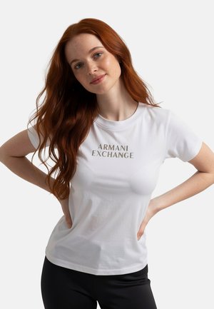 Giovane donna con lunghi capelli rossi che indossa una maglietta bianca Armani Exchange e pantaloni neri, posa con le mani sui fianchi, sorridendo dolcemente.