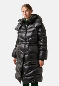 Black puffer coat with a glossy finish, featuring a high collar, zip closure, and oversized, quilted design. Long sleeves and pockets included.