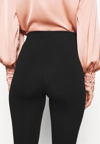 Zwarte high-waisted leggings met een gladde textuur, gecombineerd met een roze satijnen blouse met geplooide mouwen en knoopaccenten.