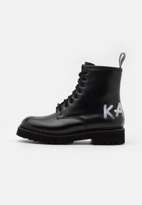 Botín de cuero negro hasta el tobillo con diseño de cordones, suela gruesa y detalle en estilo graffiti "KA" en el lateral. Cuenta con una pestaña en la parte superior para facilitar su puesta.