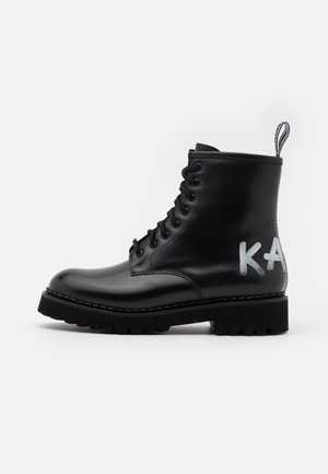 Stivaletto in pelle nera con design a lacci, suola spessa e dettaglio in stile graffiti "KA" sul lato. Presenta una linguetta per una facile calzata.