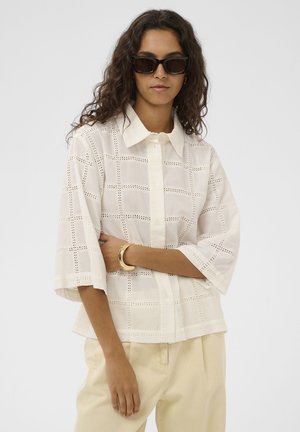 Kehar lokki ja mustad päikeseprillid, valge pastapallist pitsist nööbitav särk laiade varrukatega, beežid püksid ja kuldne käevõru.