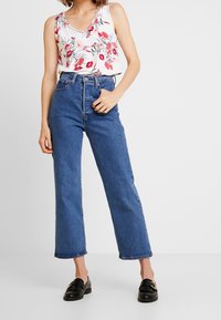 Donna che indossa un top floreale bianco senza maniche, jeans blu a vita alta e mocassini neri, in piedi contro uno sfondo bianco semplice.