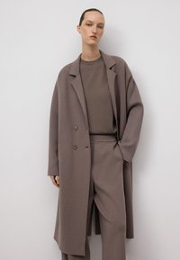 Donna che indossa un cappotto oversize mauve, pantaloni larghi abbinati e una maglietta a girocollo, in piedi contro uno sfondo bianco semplice.