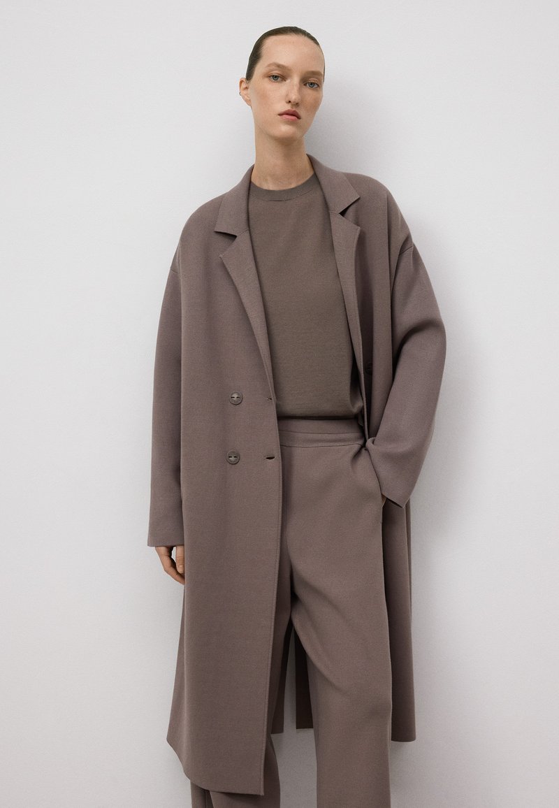 Donna che indossa un cappotto oversize mauve, pantaloni larghi abbinati e una maglietta a girocollo, in piedi contro uno sfondo bianco semplice.