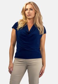 Granatowa bluzka z dekoltem w kapturek, wykonana z gładkiego materiału, z krótkimi rękawami, zestawiona z jasnobeżowymi dopasowanymi spodniami, prezentowana na neutralnym tle.