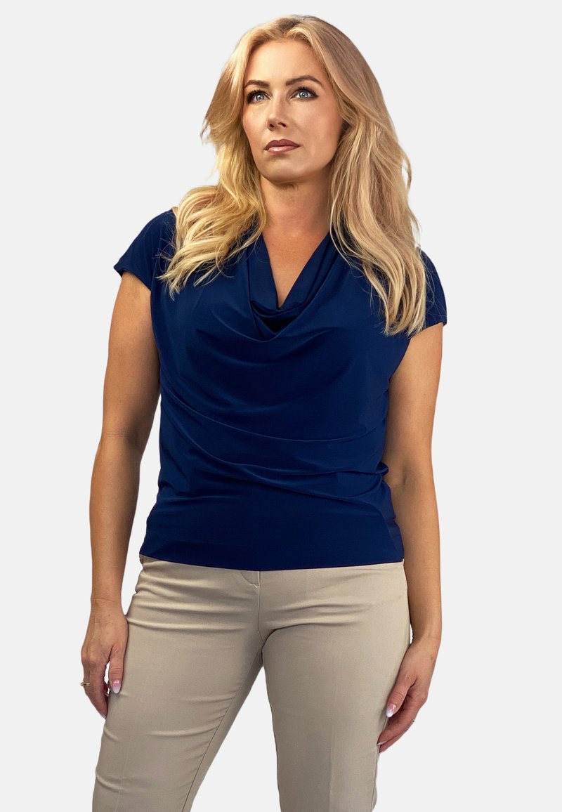 Granatowa bluzka z dekoltem w kapturek, wykonana z gładkiego materiału, z krótkimi rękawami, zestawiona z jasnobeżowymi dopasowanymi spodniami, prezentowana na neutralnym tle.