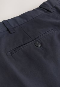 Pantalon en coton bleu marine avec une texture lisse, doté d'une poche arrière avec un bouton noir et d'une passants pour ceinture. Design simple et minimaliste.