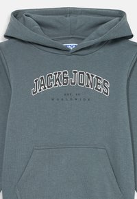 Hanorac cu glugă din material verde teal închis, prevăzut cu buzunar frontal și text alb cu inscripția „JACK&JONES” și „EST. 90 WORLDWIDE” dedesubt.