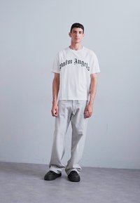 Hvit T-skjorte med svart "Palm Angels" tekst, kombinert med lys grå jeans og svarte joggesko, stående mot en nøytral bakgrunn.