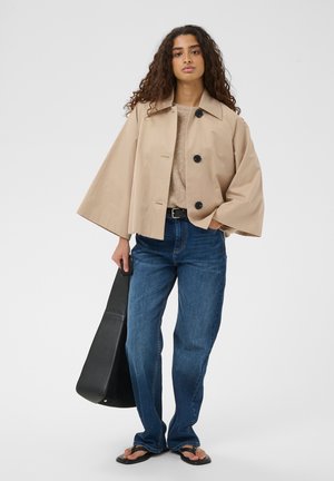 Kiharatega naine, kes kannab beežikat manteljakki, helepruuni kampsunit, siniseid teksaseid ja musti varbavahekingi, hoides suurt musta käekotti valgel taustal.