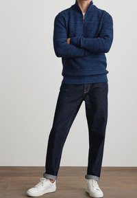 Marineblauer Strickpullover mit einem Reißverschlusskragen, kombiniert mit dunklen Jeans und weißen Sneakers. Strukturiertes Material mit gerippten Bündchen.