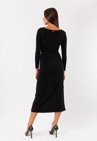 Abito nero a maniche lunghe con forma aderente, lunghezza al ginocchio, texture liscia e un sottile dettaglio sul retro. Abbinato a tacchi alti neri.