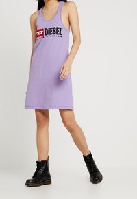 Ärmlös lavendelfärgad klänning med en svart "DIESEL"-logotyp och texten "DENIM DIVISION", tillverkad av mjukt material. Kombinerad med svarta snörstövlar.