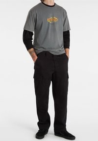 Camiseta gris con un logo colorido en el pecho, superpuesta a una camiseta de manga larga negra, combinada con pantalones cargo negros y zapatos negros.