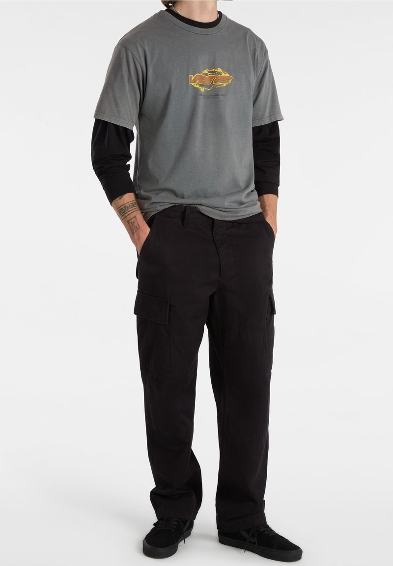 Camiseta gris con un logo colorido en el pecho, superpuesta a una camiseta de manga larga negra, combinada con pantalones cargo negros y zapatos negros.