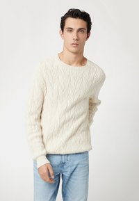Maglione a trecce color crema con un classico scollo rotondo. La trama presenta un motivo a onda, abbinato a jeans in denim blu chiaro.