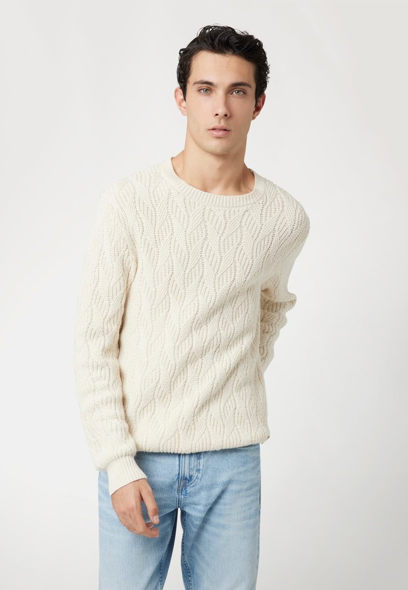 Maglione a trecce color crema con un classico scollo rotondo. La trama presenta un motivo a onda, abbinato a jeans in denim blu chiaro.