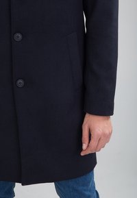 Cappotto di lana blu navy con una texture liscia, due bottoni e tasche laterali, abbinato a jeans di denim blu.