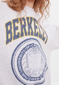 Grå sweatshirt med "BERKELEY" i gult och marinblått, med en universitetssegelgrafik som föreställer en bok och ett band nedan.