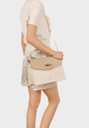 Donna che indossa una maglietta beige a maniche corte e una gonna floreale, che tiene una borsa beige e crema con una fibbia intrecciata dorata.