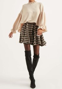Beige top s dlouhým rukávem a volánkovým výstřihem, doplněný černou a béžovou sukní s kostkovaným vzorem a černými kozačkami po kolena.