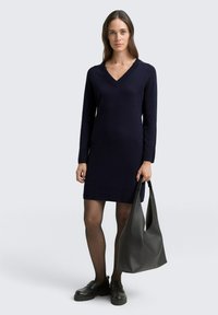 Granatowa dzianinowa sukienka z dekoltem w serek i długimi rękawami, zestawiona z gładką czarną torbą na ramię i masywnymi czarnymi loafersami. Look jest prosty i schludny.