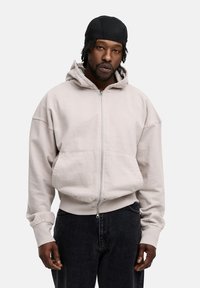 Light gray zip-up hoodie with a loose fit, large front pockets, and a drawstring hood, paired with black jeans. Soft cotton material.