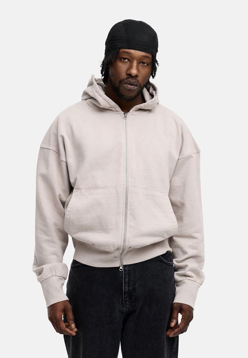 Light gray zip-up hoodie with a loose fit, large front pockets, and a drawstring hood, paired with black jeans. Soft cotton material.