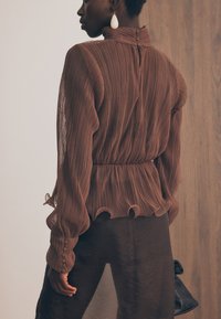 Brūna krokotā blūze ar augstu kakla izgriezumu un pogu aizdari aizmugurē, ar riņķotu apakšmalu un garām piedurknēm ar pogu akcentiem.