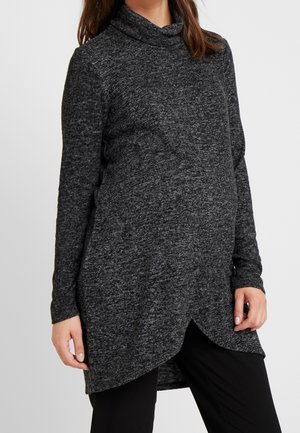 Tunique à manches longues noire et grise chinée avec un col cowl, un ourlet asymétrique et une texture tricot douce, portée sur un pantalon noir.