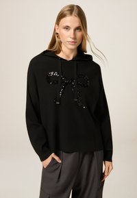 Schwarzer Kapuzenpullover aus weichem Stoff mit einem großen Paillettenbogen-Design auf der Vorderseite, mit Kordelzügen und einer lässigen Passform.