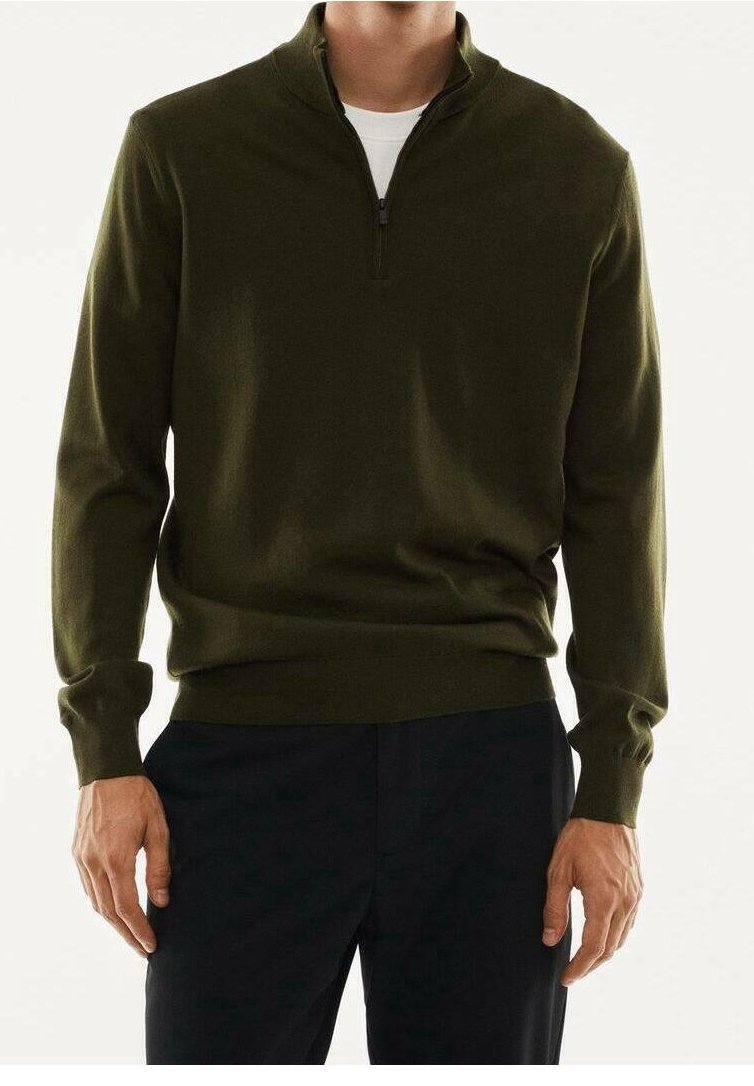 Homme portant un pull vert olive à demi-fermeture éclair sur une chemise blanche et un pantalon foncé, debout avec les bras détendus le long du corps.
