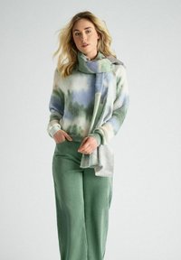 Zachte, gebreide trui met een blauw-groene gradient patroon, gecombineerd met een gestructureerde groene wijde broek en een lichte, patroon sjaal.