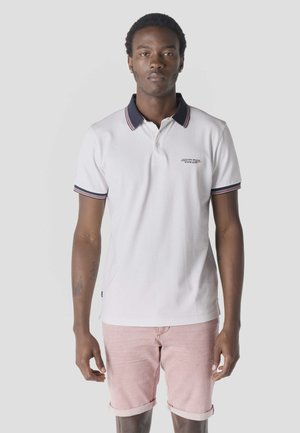 Joven vestido con una camiseta polo blanca con cuello azul marino y pantalones cortos de color rosa claro remangados, de pie frente a un fondo gris liso.