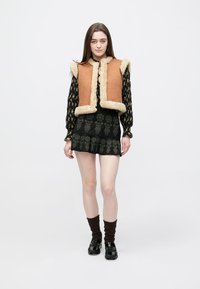 Καφέ σουέντ γιλέκο με φόδρα από κρεμ συνθετική γούνα, φορεμένο πάνω από μαύρο φόρεμα με μοτίβα και βολάν στο τελείωμα. Μαύρα παπούτσια με πλατφόρμα και σκούρες κάλτσες.