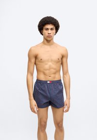 Μπόξερ σε ναυτικό μπλε χρώμα με κάθετες κόκκινες και λευκές ρίγες, ελαστική μέση και μικρή ετικέτα μάρκας στο μπροστινό μέρος. Ομαλή υφή υφάσματος.