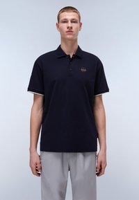 Marinblå polo-shirt med klassisk krage, tre knappar och en liten rund logo. Kortärmad med kontrasterande kantdetalj. Kombinerad med ljusgrå byxor.