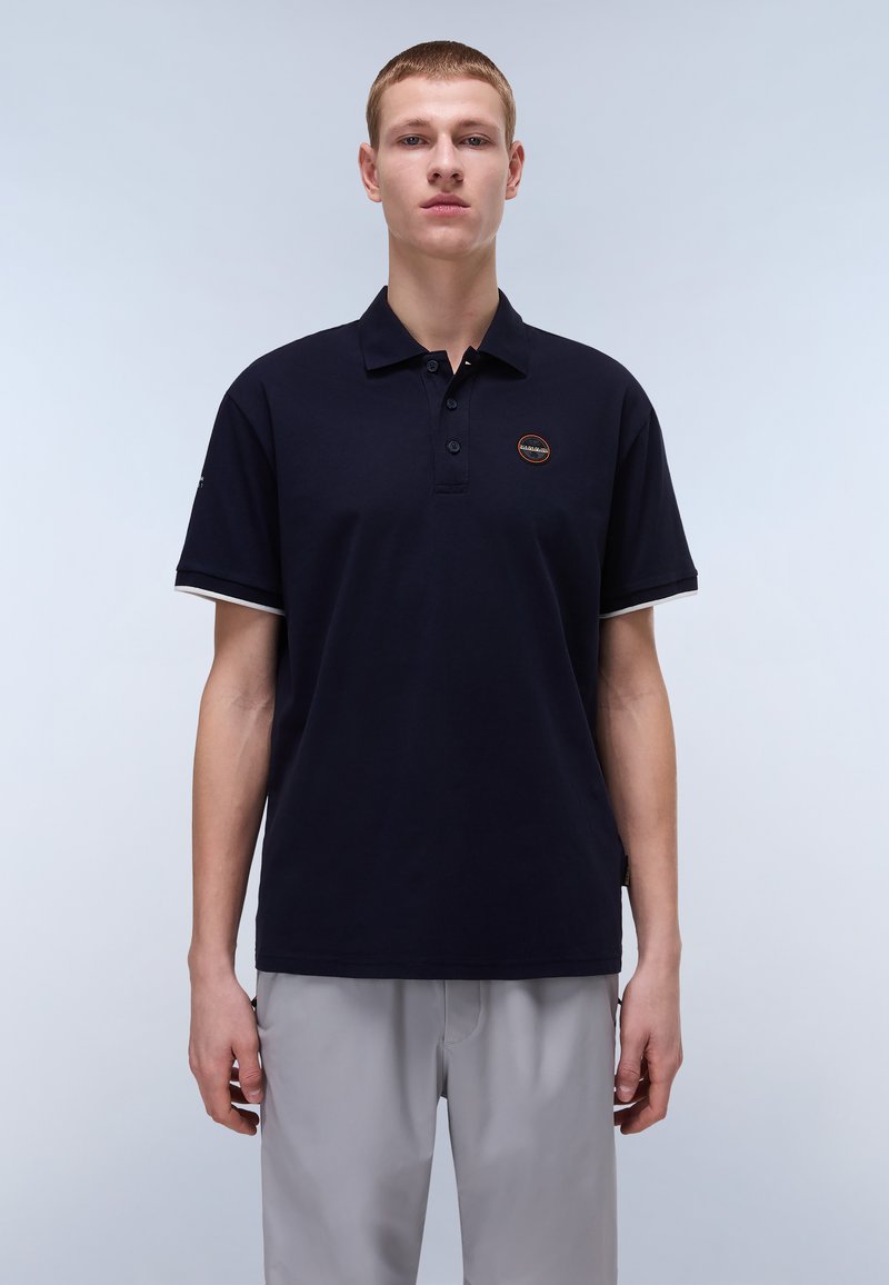 Marinblå polo-shirt med klassisk krage, tre knappar och en liten rund logo. Kortärmad med kontrasterande kantdetalj. Kombinerad med ljusgrå byxor.