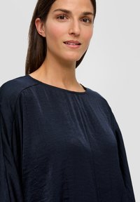 Marineblaues Langarmoberteil mit rundem Ausschnitt, aus einem glatten, leichten Stoff gefertigt. Besitzt einen subtilen Glanz und eine lockere Passform.