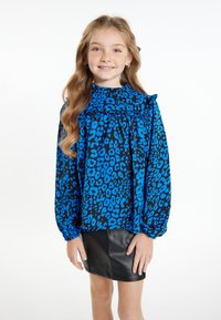 Blå leopardtrykt bluse med flæser ved halsen og lange ærmer, parret med en sort lædernederdel. Glat stof og livlige farver.