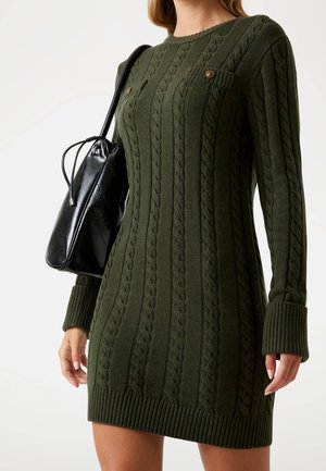 Vrouw die een donker groen kabelgebreide sweaterjurk draagt met omgeslagen mouwen en met knopen sluitende borstzakken, en een zwarte leren schoudertas draagt.