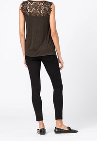 Kvinna som bär en svart ärmlös topp med spetsbak, svarta tight jeans och svarta texturerade platta skor, stående mot vit bakgrund.