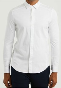 Biała, długa elegancka koszula z guzikami i klasycznym kołnierzykiem, o gładkiej fakturze i subtelnym haftowanym detalu na piersi. Połączona z ciemnymi spodniami.