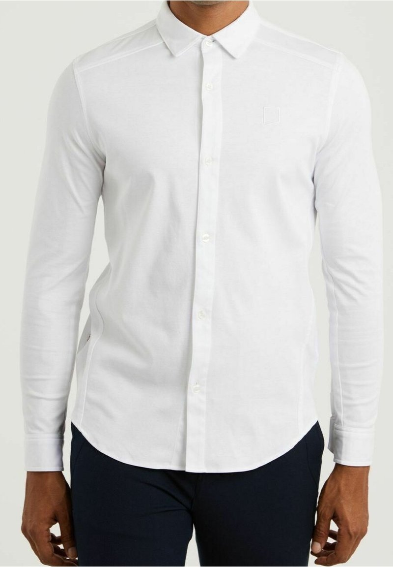 Biała, długa elegancka koszula z guzikami i klasycznym kołnierzykiem, o gładkiej fakturze i subtelnym haftowanym detalu na piersi. Połączona z ciemnymi spodniami.