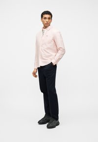 Homme portant une chemise rose clair à boutons par-dessus un t-shirt rayé, un pantalon bleu marine et des baskets noires, debout devant un fond blanc uni.
