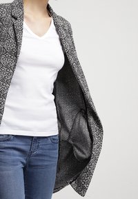 Svart och vit mönstrad kavaj med en texturerad tyg, öppen framtill, bärs över en vit V-ringad t-shirt och blå jeans.